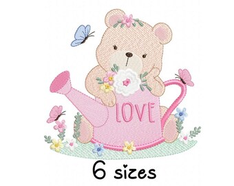 May include: Embroidered design of a brown bear holding a pink watering can with the word "LOVE" on it. The bear is wearing a flower crown and surrounded by flowers and butterflies.