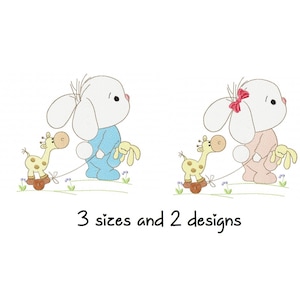 Pode incluir: Dois desenhos bordados de um coelho branco segurando um brinquedo de girafa. Um coelho está vestindo azul e o outro rosa. O texto "3 tamanhos e 2 designs" está abaixo dos coelhos.