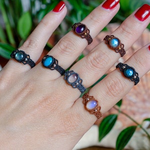 Op de afbeelding: Een hand draagt zes ringen met verschillende gekleurde edelstenen. De ringen zijn gemaakt van bruin en zwart koord en hebben kleine gouden kralen. De edelstenen zijn amethyst, maansteen, turquoise, rozekwarts en labradoriet.