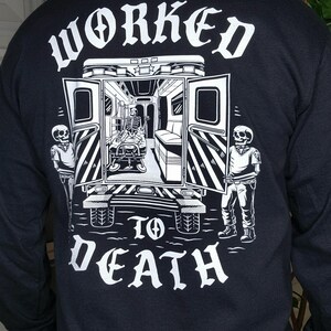 May include: Black long-sleeve shirt with a graphic on the back. The graphic features a skeleton in an ambulance with the words "WORKED TO DEATH" in white gothic lettering. Two skeleton figures stand on either side of the ambulance.