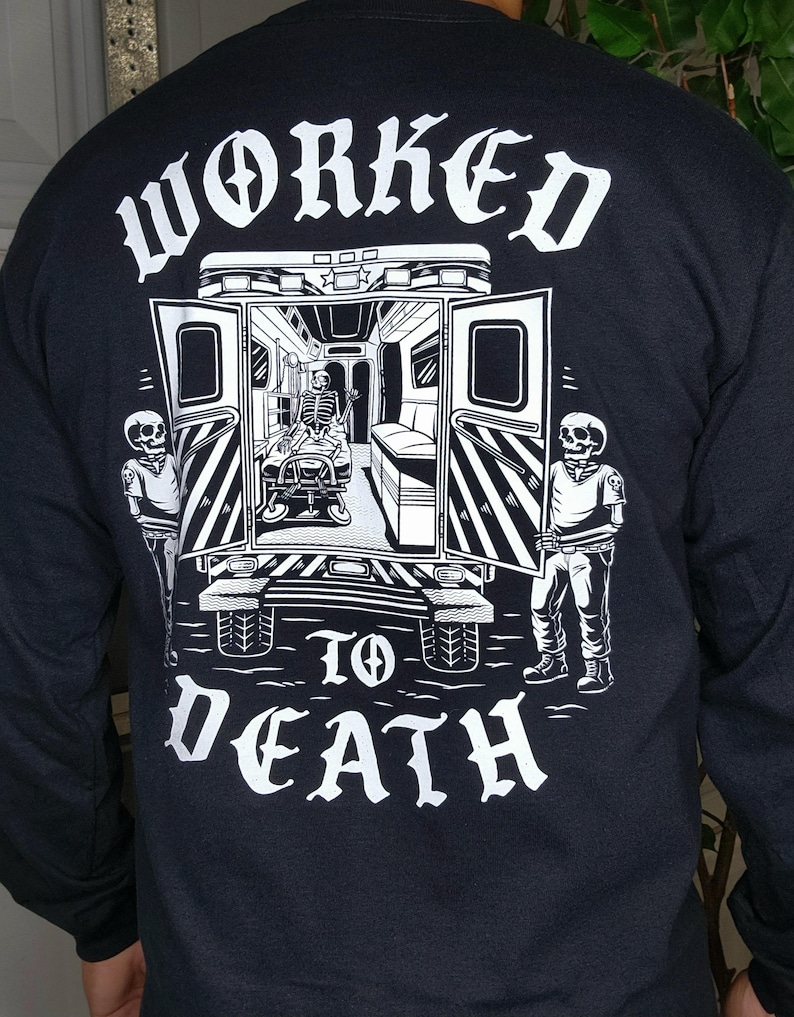 May include: Black long-sleeve shirt with a graphic on the back. The graphic features a skeleton in an ambulance with the words "WORKED TO DEATH" in white gothic lettering. Two skeleton figures stand on either side of the ambulance.
