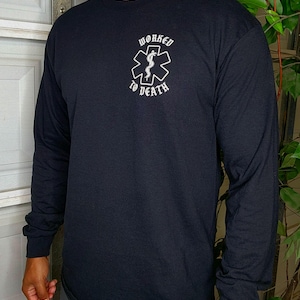 May include: Black long-sleeve shirt with the text "WORKED TO DEATH" above a white medical symbol. The shirt is made of a soft material and has long sleeves. The design is centered on the chest.