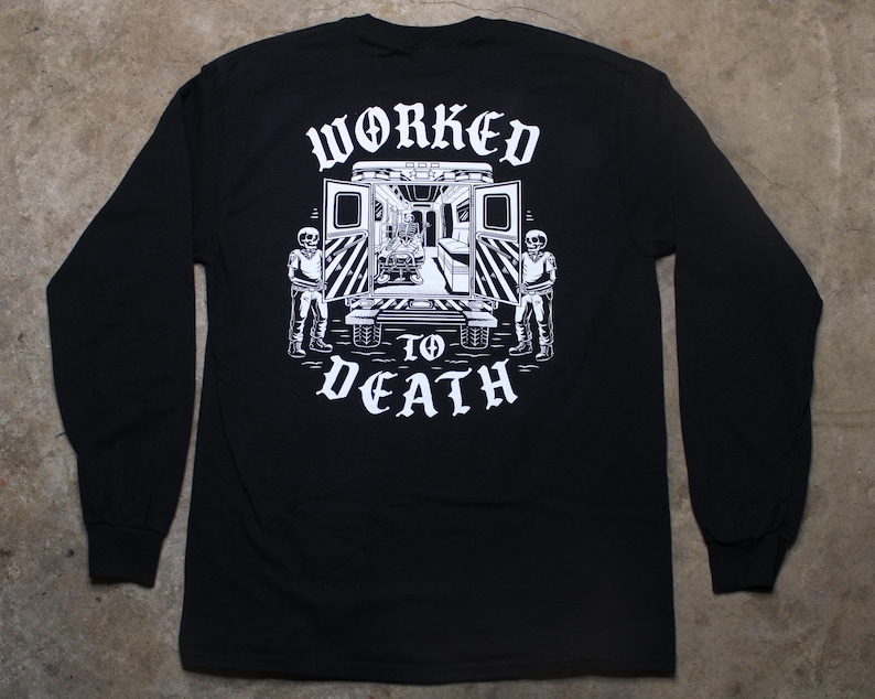 May include: Black long-sleeve shirt with a white graphic on the back. The graphic features an ambulance with skeletons and the words "WORKED TO DEATH" in a gothic font. The shirt has a crew neck and long sleeves.