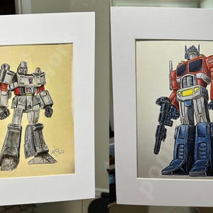 Pode incluir: Duas ilustrações emolduradas de personagens Transformers. O quadro da esquerda apresenta Megatron, um robô cinza com uma grande arma. O quadro da direita apresenta Optimus Prime, um robô vermelho, azul e branco com uma arma.
