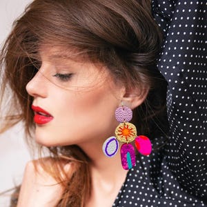 May include: A woman wearing a black and white polka dot shirt with colorful geometric earrings. The earrings are made of multiple shapes and colors, including pink, orange, yellow, green, and purple.
