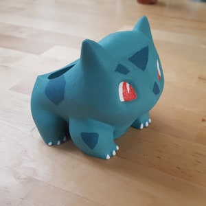 Peut inclure: Une figurine en 3D imprimée en bleu turquoise représentant un personnage Pokémon, Bulbizarre. La figurine a des yeux rouges et des accents blancs.