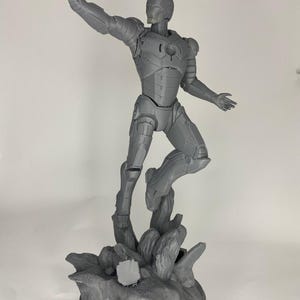Può includere: Una statuetta di Iron Man grigia, stampata in 3D, in posa dinamica, con un braccio alzato e l'altro teso. La statuetta è montata su una base rocciosa. La statuetta è una rappresentazione dettagliata dell'armatura del supereroe.