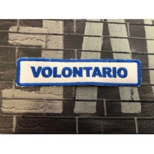 Può includere: Toppa rettangolare con la scritta "VOLONTARIO" ricamata in blu su sfondo bianco, bordata di blu. La toppa è posta su un muro di mattoni strutturato.