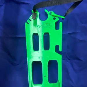 Può includere: Un attrezzo verde brillante, rettangolare, con più sezioni ritagliate e un cinturino nero. Il testo "infostudio" è stampato. L'attrezzo ha una serie di tacche lungo il bordo superiore.