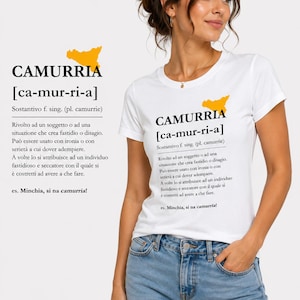 Può includere: Una t-shirt bianca a maniche corte con una stampa grafica della parola CAMURRIA, la sua pronuncia fonetica, una definizione in italiano e una piccola mappa gialla della Sicilia. La maglietta è abbinata a un paio di jeans azzurri.