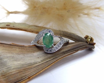May include: A silver ring with an oval, emerald-green gemstone at the center, surrounded by small, clear accent stones. The ring's band curves in a swirling design. The ring is displayed on a brown, textured surface.