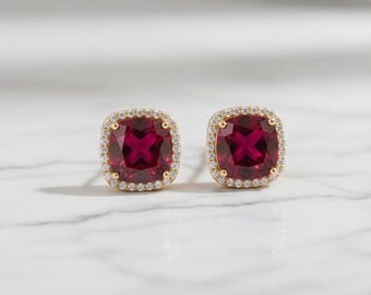 May include: A pair of gold-toned stud earrings. Each earring features a square, deep red gemstone surrounded by a halo of small, clear crystals. The earrings are set against a white marble surface.