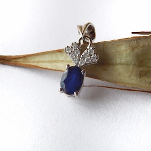 May include: A silver pendant featuring a deep blue, oval-cut gemstone. The pendant is accented with smaller, clear gemstones and is suspended from a delicate silver bail. The design is elegant and suitable for everyday wear or special occasions.