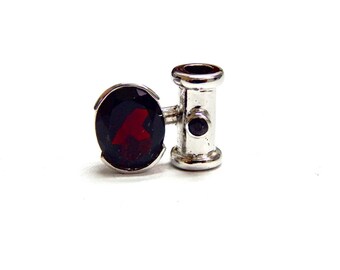May include: A silver-colored charm with a dark red oval-shaped stone featuring a red letter "A". The charm has a cylindrical design with a small, dark stone on the side. The charm is set against a white background.
