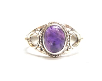 May include: A silver ring featuring an oval, deep purple gemstone. The ring has a decorative, rope-like border around the gemstone and openwork designs on either side. The ring is set against a white background.