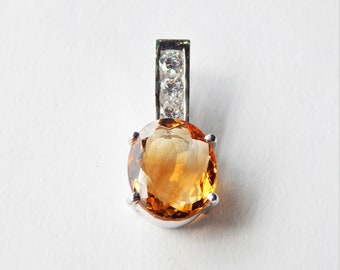 May include: A silver pendant featuring an oval, amber-colored gemstone. The gemstone is set in a silver bezel and is topped with a rectangular bail adorned with three small, clear stones. The pendant is displayed against a white background.