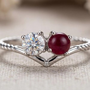 Può includere: Un anello color argento con un design a fascia intrecciata che converge a forma di V. L'incastonatura contiene una pietra rotonda sfaccettata trasparente e una pietra rotonda liscia rosso scuro affiancate, con una minuscola pietra alla base della V.