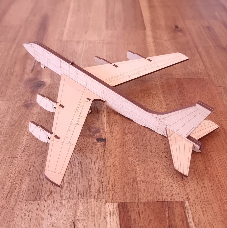 May include: A wooden model of a B-52 bomber airplane. The model is made of light brown wood and has detailed markings. The model is lying on a wooden surface.