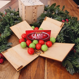 Może przedstawiać: Drewniane pudełko z wycięciami w kształcie gwiazd i otworem w kształcie gwiazdy, otoczone świąteczną girlandą. Pudełko jest otwarte, ukazując czerwoną i białą kartę z napisem "CLUB+" oraz czerwone i zielone cukierki.