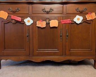 May include: A brown wooden cabinet with three drawers and three doors. A string of felt breakfast food cutouts hangs above the cabinet. The cutouts include a pancake, a fried egg, bacon, and toast.