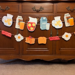 May include: A breakfast-themed banner with various food items, including orange juice, milk cartons, bacon, eggs, pancakes, and a pitcher of orange juice. The banner is strung with twine and hung on a wooden cabinet.