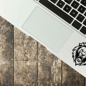 May include: A black and white lion head sticker is affixed to the corner of a laptop. The laptop is on a weathered, wooden surface. The lion has an open mouth and a fierce expression.