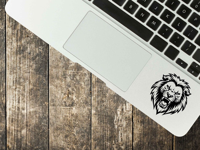 May include: A black and white lion head sticker is affixed to the corner of a laptop. The laptop is on a weathered, wooden surface. The lion has an open mouth and a fierce expression.