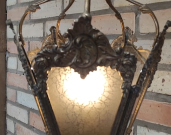 May include: A hanging lantern pendant light featuring an ornate, antique brass-toned metal frame with intricate scrollwork and floral detailing. The fixture contains frosted glass panels with a crackled texture, illuminated from within by a light bulb.