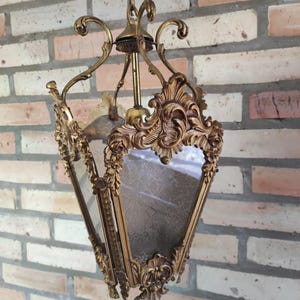 Puede incluir: Lámpara colgante dorada ornamentada con un marco decorativo y paneles espejados. La lámpara presenta intrincados detalles de volutas y florales, suspendida por una cadena. La lámpara de estilo vintage está contra una pared de ladrillos.
