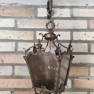 Puede incluir: Lámpara colgante de estilo antiguo con acabado bronce. La lámpara presenta una parte superior decorativa, intrincados trabajos en metal y paneles de vidrio texturizado. Está suspendida de una cadena y montada contra una pared de ladrillo.