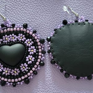Könnte beinhalten: Ein Paar runde Ohrringe mit einem zentralen schwarzen Herz-Cabochon, umgeben von kleinen lavendelfarbenen und schwarzen Saatperlen in einem Blumenmuster. Die Ränder sind mit schwarzen Perlen akzentuiert und die Rückseite zeigt einen glatten schwarzen Untergrund.