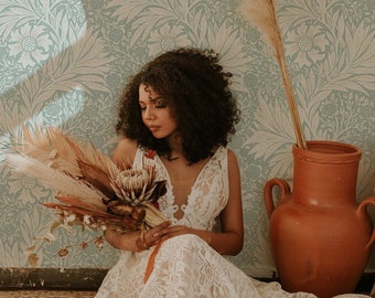 May include: A woman in a white lace dress sits on a rug in front of a wall with a floral pattern. She is holding a bouquet of dried flowers and pampas grass. There is a terracotta pot on the right side of the image.