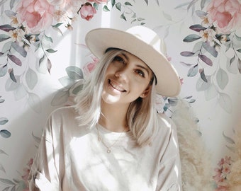 May include: A woman wearing a white hat and a light beige t-shirt sits in front of a floral wallpaper. She is holding a white mug with the text "James Coffee Co." printed on it.