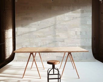 May include: A wooden table on sawhorses with a black metal stool in front of a white brick wall. The table and stool are in a white room with natural light.