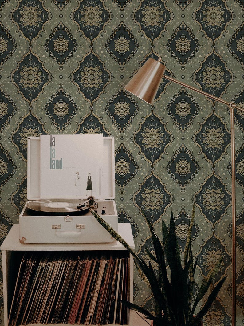 May include: A record player with a vintage record on a shelf with a stack of records. The record player is in front of a wall with a dark green and gold patterned wallpaper. A plant and a lamp are also in the image.