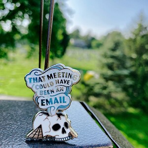 May include: A silver enamel pin with a white skull and a blue cloud with the text "That meeting could have been an email".