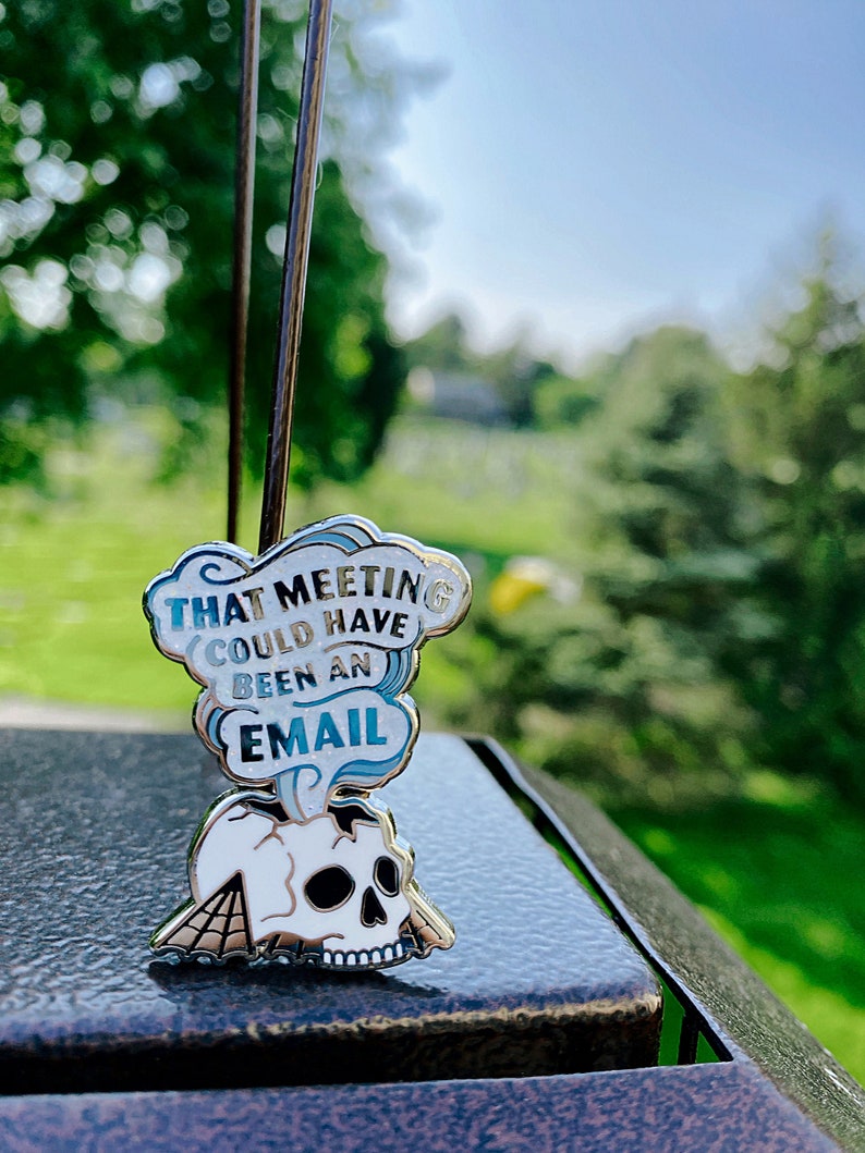 May include: A silver enamel pin with a white skull and a blue cloud with the text "That meeting could have been an email".