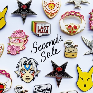 May include: A collection of enamel pins featuring various designs, including a star with a demon face, a heart with a crown, a cartoon dumpling, and a character from a video game. The pins are arranged on a white background with the text "Seconds Sale" written in black.