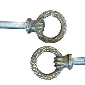 May include: Two antique brass key pulls with a decorative design. Each pull features a circular handle with a detailed floral pattern and a hand-shaped grip. The pulls have a silver-colored stem.