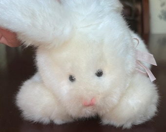 May include: A plush white rabbit toy with soft fur, dark eyes, and a small pink nose. The stuffed animal features a light pink ribbon tied in a bow near the ear, with one ear positioned upright and the other folded down against the head.