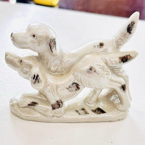 May include: A ceramic figurine of two dogs in a playful pose. The dogs are off-white with brown spots and are set on a matching base. The figurine is likely a decorative item.