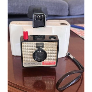 May include: A vintage Polaroid Swinger Model 20 land camera. The camera has a black body with a gold-colored front and a large lens. The camera has a red button and a black strap. A white flash attachment is behind the camera.