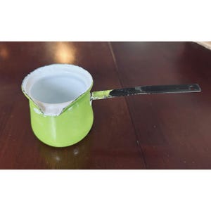Puede incluir: Una pequeña cafetera de esmalte verde lima con un mango largo y negro. El interior es blanco y la cafetera tiene un pico para verter fácilmente. El mango tiene un agujero para colgar. Un artículo de cocina vintage.