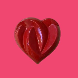 May include: A glossy, heart-shaped object in a vibrant red hue. The heart features a unique, interwoven design with curved lines, set against a bright pink background. The object has a smooth, reflective surface.