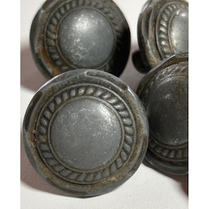 May include: A collection of round, dark gray metal cabinet knobs featuring a smooth, raised center dome surrounded by a textured, rope-like border. The surface of each knob displays a weathered, matte finish with subtle signs of oxidation.