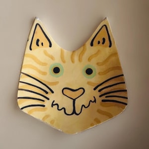May include: A whimsical, hand-painted ceramic cat face with a yellow and orange tabby pattern. The cat has green eyes, black whiskers, and a smiling mouth. The ears are pointed, and the overall design is playful and decorative.