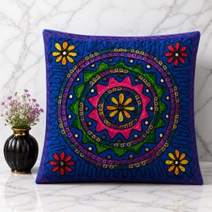 May include: A square blue throw pillow featuring a central circular mandala pattern embroidered with vibrant pink, green, and yellow thread, accented with small reflective mirror pieces and four floral motifs in the corners.