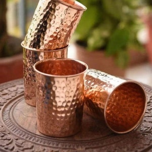 Puede incluir: Varios vasos de color cobre con una textura martillada. Los vasos son cilíndricos con un borde ligeramente ensanchado. Algunos están apilados, otros están tumbados de lado sobre una superficie de madera. Los vasos tienen un brillo metálico cálido.