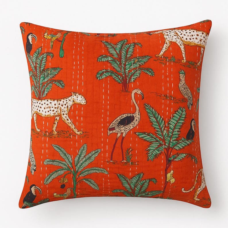 May include: Square orange pillow with a repeating pattern of leopards, flamingos, toucans, and palm trees. The design is stitched with white thread. The pillow has a tropical theme.