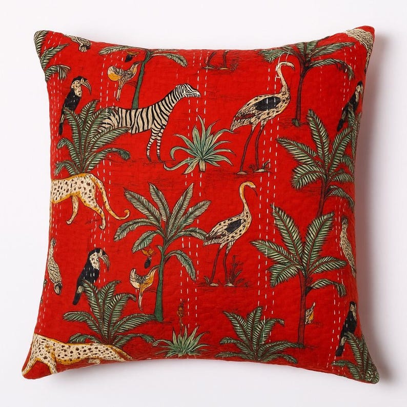 May include: Red square throw pillow with a vibrant jungle scene. The pillow features a zebra, leopard, birds, and palm trees. The design is printed on the fabric and stitched with white thread. The pillow has a solid red background.